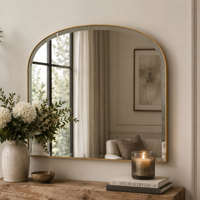 An arched wall mirror with a thin gold frame hangs above a rustic wooden console table. The mirror reflects a softly lit interior with curtains, a framed print, and a chair, while the room itself is styled with a large white flower arrangement in a textured vase and a lit candle on stacked books.