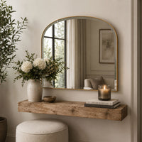 An arched wall mirror with a gold frame hangs above a rustic wooden console shelf in a softly styled interior. A vase of white flowers, a candle, books, and a round pouf are arranged below, with the mirror reflecting a softly lit room.