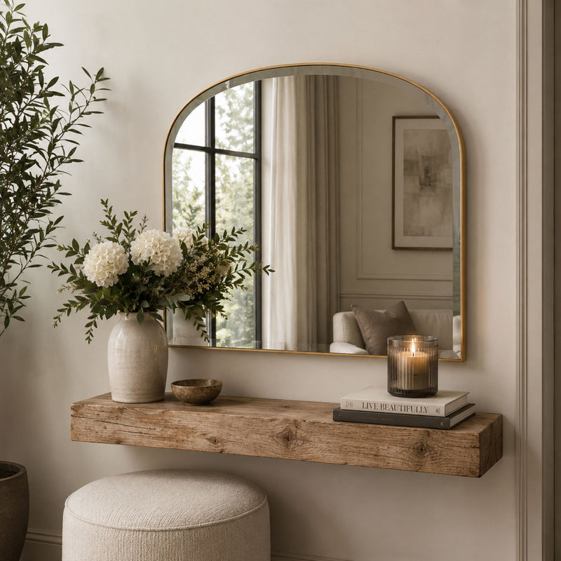 An arched wall mirror with a gold frame hangs above a rustic wooden console shelf in a softly styled interior. A vase of white flowers, a candle, books, and a round pouf are arranged below, with the mirror reflecting a softly lit room.