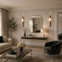 A styled interior room with cream paneled walls, a large oval wall mirror, wall sconces, a console table, a sofa, a dark green velvet armchair, a marble coffee table, and decorative plants and vases.