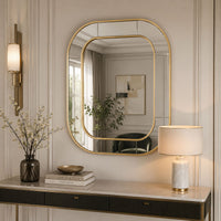 An elegant gold-framed wall mirror with an asymmetrical rounded top, styled in a classic cream-colored room above a console table with a lamp and books.