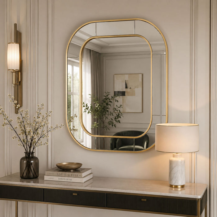 An elegant gold-framed wall mirror with an asymmetrical rounded top, styled in a classic cream-colored room above a console table with a lamp and books.