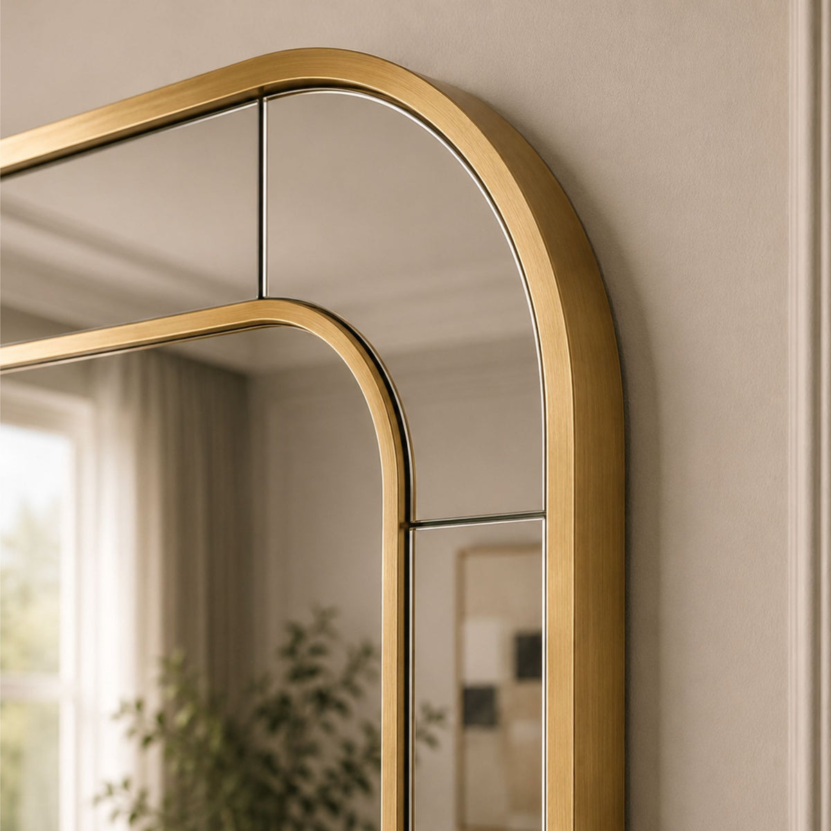 A close-up of a modern wall mirror with a brushed gold arched frame, mounted on a beige wall with a soft reflection of a room interior visible.