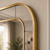 A close-up of a modern wall mirror with a brushed gold arched frame, mounted on a beige wall with a soft reflection of a room interior visible.