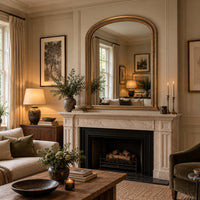 A large arched gold-framed mirror hangs above a white marble fireplace mantel in a beautifully styled living room with beige walls, a wooden coffee table, and soft warm lighting.