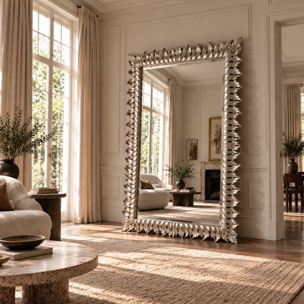 A large floor mirror with an ornate silver frame stands in a bright, elegant living room with cream walls, tall windows, and natural light.
