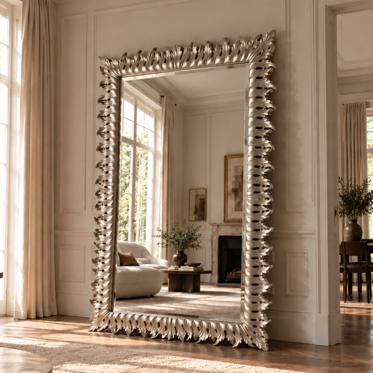 A large floor-length mirror with an ornate, sculptural silver frame stands in a classic cream-colored room with wood floors and window light.