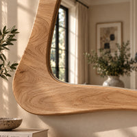 A large, uniquely shaped wooden wall mirror with a curved, organic frame, shown mounted on a beige wall in a beautifully lit room with soft sunlight and blurred interior decor.