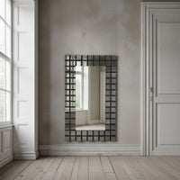 A rectangular wall mirror with a dark, geometric grid frame hangs on a textured gray wall in a bright, modern room with white paneled doors and light wood floors.