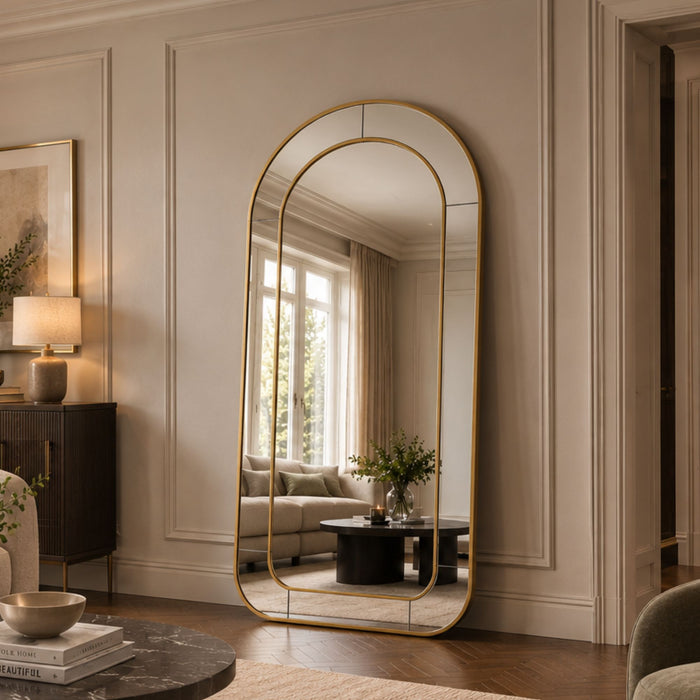 A tall, arched floor mirror with a thin gold frame stands in a styled living room with cream paneled walls, a sofa, and a coffee table.