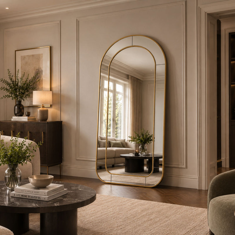 A tall arched full-length floor mirror with a thin gold frame stands in a styled living room with cream paneled walls, a dark wood console, and a sofa reflected in the mirror.
