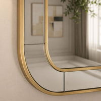 An arched wall mirror with a brushed gold metal frame, shown mounted on a beige wall with its reflection visible.