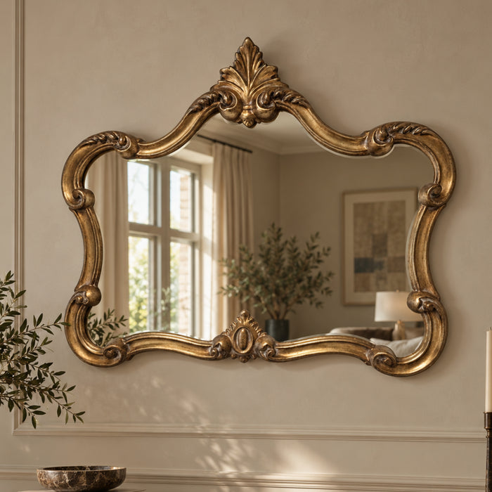 An ornate gold-framed wall mirror with a decorative baroque-style frame, mounted on a beige wall. The mirror reflects a softly lit room with a window, curtains, and a plant.