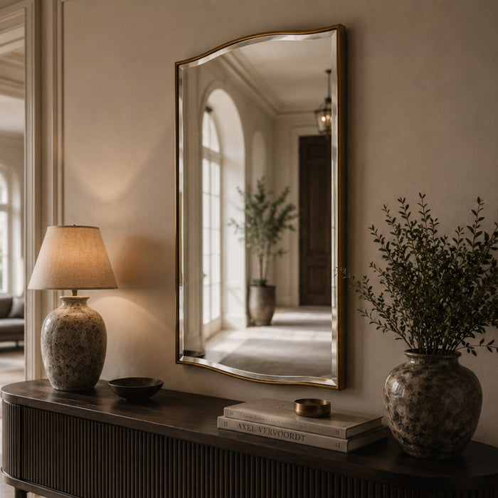 A tall, arched wall mirror with a gold-toned frame hangs on a beige wall above a dark wood console table. The mirror reflects a softly lit hallway with a potted plant, while the table is styled with a lit table lamp, books, and a large ceramic vase with greenery.