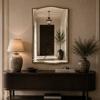 A large arched wall mirror with a gold frame hangs above a dark wood console table in a warm, elegant room. The mirror reflects a dark wooden door and a hanging lantern, while the room itself is styled with a patterned ceramic table lamp on the left and a matching vase with dried greenery on the right.