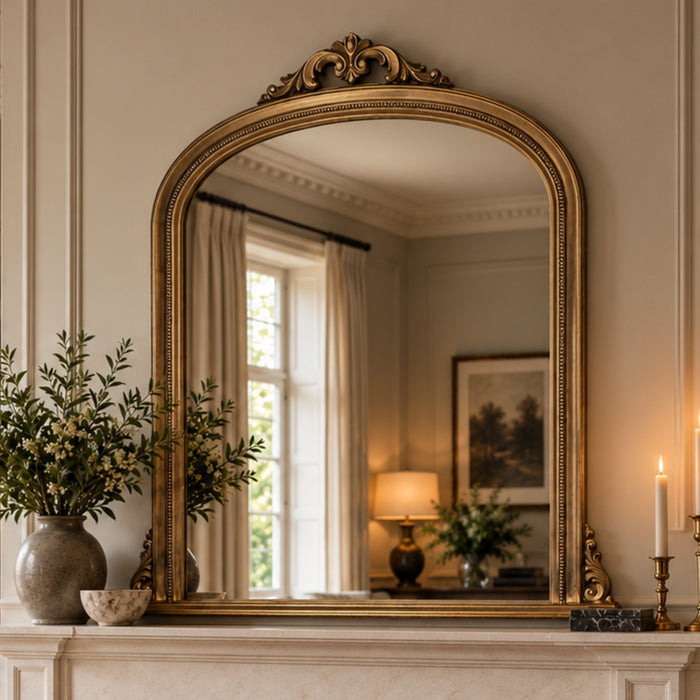 An ornate gold-framed arched mirror with a decorative crest, displayed on a white fireplace mantel in a warm, elegant room setting.