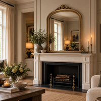An ornate gold-framed mirror with a curved top is mounted above a white fireplace mantel in a beautifully styled living room with warm lighting, candles, and floral arrangements.