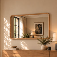 A rectangular wall mirror with a light natural wood frame hangs above a matching wooden dresser in a warm, sunlit room. The mirror reflects a window with curtains and framed wall art, while the dresser surface holds books, a bowl, a vase with greenery, and a lamp.