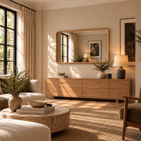 A warm, modern living room featuring a light wood console cabinet with a large matching wood-framed mirror, styled with a table lamp, ceramic vases, and plants. The room has beige walls, a textured woven rug, and natural sunlight streaming through a window with black frames.