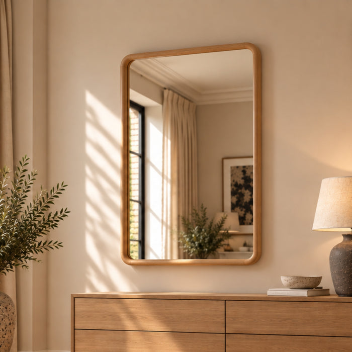 A rectangular wall mirror with a light natural wood frame hangs on a beige wall above a matching wood dresser in a softly lit room. The mirror reflects a window, curtains, and indoor decor, while a lamp and plant sit on the dresser beside it.