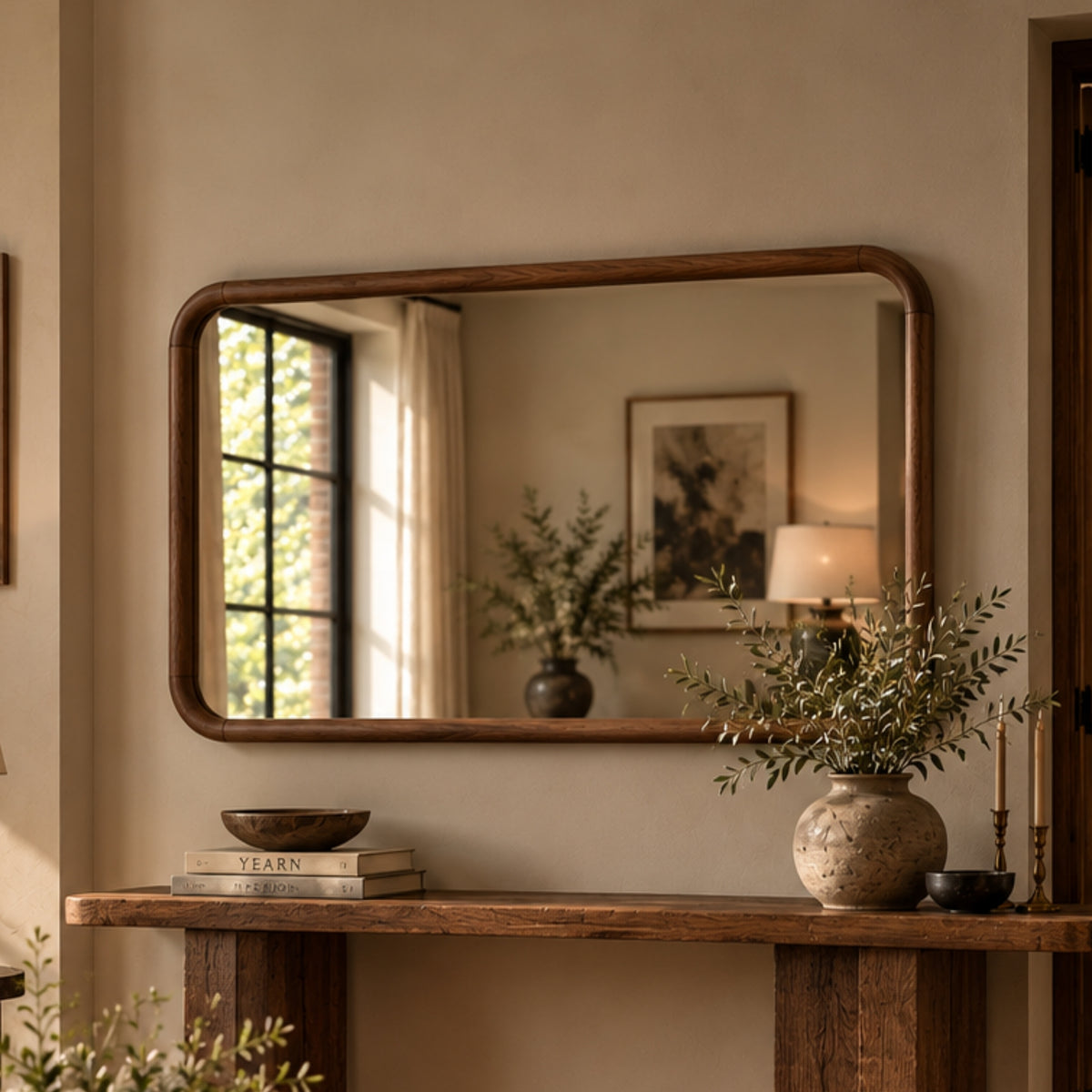 A rectangular wall mirror with a warm medium-brown wooden frame hangs above a rustic wooden console table in a styled interior. The mirror reflects a window, framed art, and a lamp, while the table holds books, a bowl, and a textured vase with greenery.