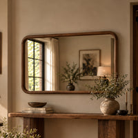 A rectangular wall mirror with a warm medium-brown wooden frame hangs above a rustic wooden console table in a styled interior. The mirror reflects a window, framed art, and a lamp, while the table holds books, a bowl, and a textured vase with greenery.