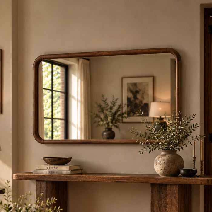 A rectangular wall mirror with a warm medium-brown wooden frame hangs above a rustic wooden console table in a styled interior. The mirror reflects a window, framed art, and a lamp, while the table holds books, a bowl, and a textured vase with greenery.