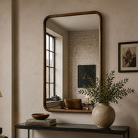 A large rectangular wall mirror with a dark wood frame hangs above a console table in a styled interior. The mirror reflects a window and room details, while a textured ceramic vase with greenery and framed wall art sit nearby on the table.