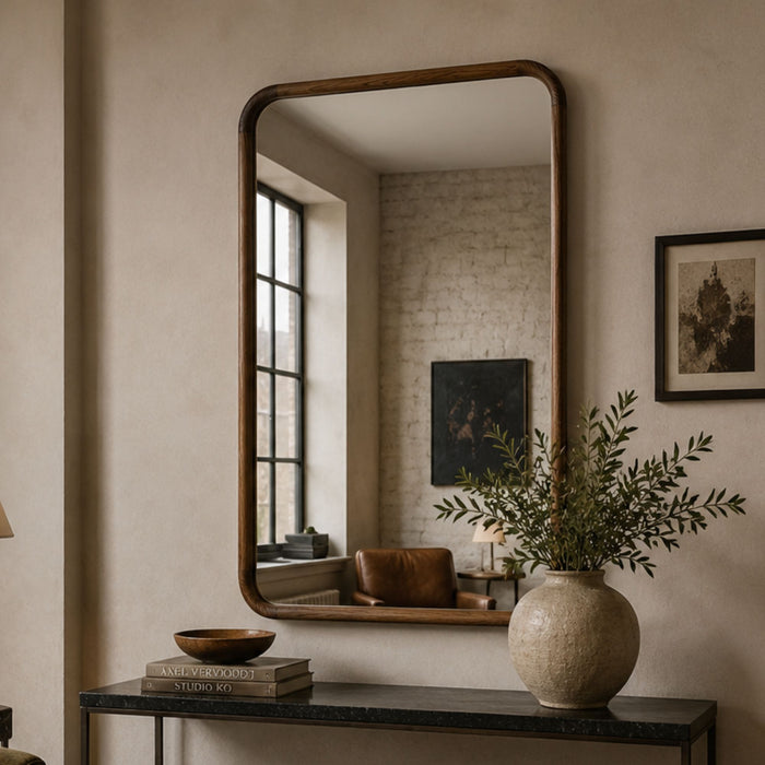 A large rectangular wall mirror with a dark wood frame hangs above a console table in a styled interior. The mirror reflects a window and room details, while a textured ceramic vase with greenery and framed wall art sit nearby on the table.