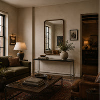 A styled living room interior with a large rectangular wall mirror, console table, lamp, sofa, coffee table, rug, and framed wall art in warm neutral tones.