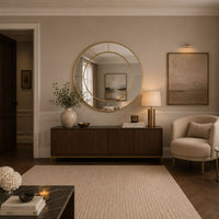 A styled interior room scene featuring a large round gold-framed mirror mounted on a beige wall above a dark wood console. The room includes a cream armchair, a textured rug, a side table with books and flowers, and framed wall art.