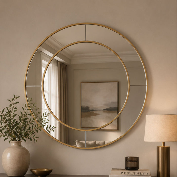 A large round wall mirror with a gold-toned double-ring frame hangs on a beige wall above a console table. The reflection shows a softly lit interior with curtains and framed art, while the room itself includes a vase with greenery, a lamp, and small decor objects.