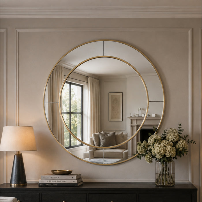 A large round wall mirror with a gold frame hangs on a beige paneled wall above a dark console table. The mirror reflects a softly lit living room with a window, curtains, and a sofa.