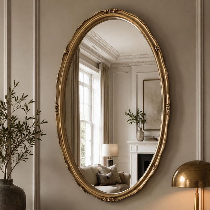 An oval wall mirror with an ornate gold frame, mounted on a beige paneled wall. The mirror reflects a softly lit interior room with a window, curtains, a sofa, and a fireplace mantel.