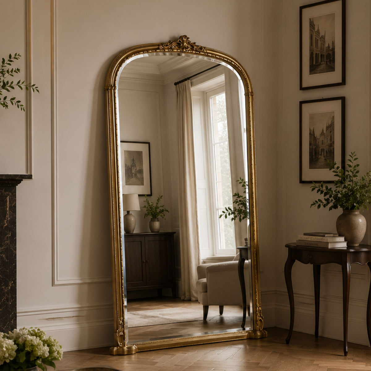 A tall floor mirror with an ornate gold frame stands in a styled room with cream walls, framed artwork, a side table, and a window reflection.