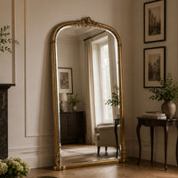 A tall floor mirror with an ornate gold frame stands in a styled room with cream walls, framed artwork, a side table, and a window reflection.