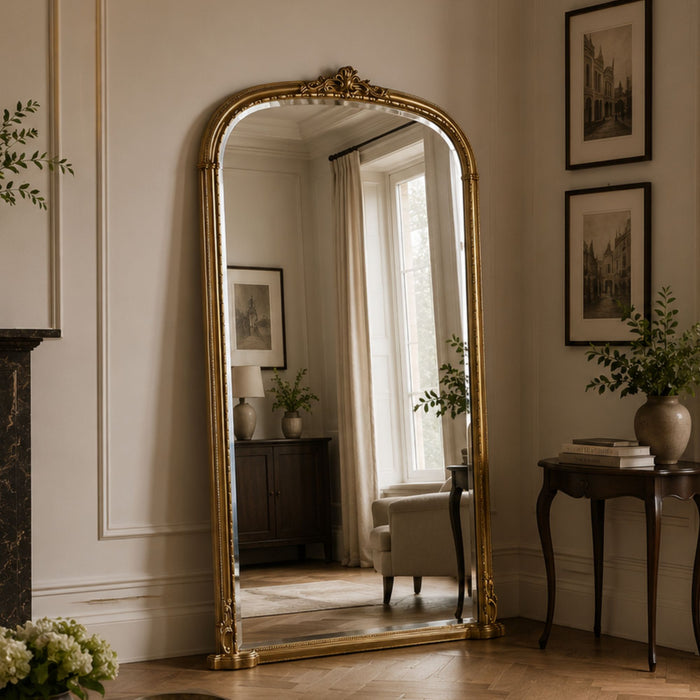A tall floor mirror with an ornate gold frame stands in a styled room with cream walls, framed artwork, a side table, and a window reflection.