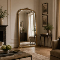 An ornate gold-framed floor mirror with an arched top stands in a classic living room with herringbone wood floors, a fireplace, and framed wall art.