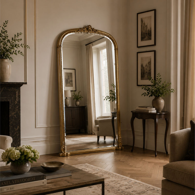 An ornate gold-framed floor mirror with an arched top stands in a classic living room with herringbone wood floors, a fireplace, and framed wall art.