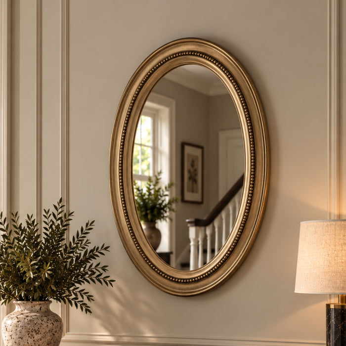 An oval wall mirror with an ornate gold frame, mounted on a beige paneled wall in a styled interior setting with a plant and lamp visible.